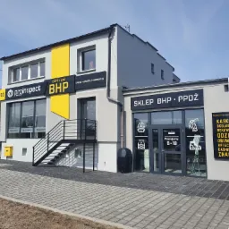 Fasada budynku firmy Proinspect BHP z napisem 'Centrum BHP', 'Usługi, Sprzedaż, Produkcja' oraz 'Sklep BHP PPOŻ', widoczne logo firmy, schody wejściowe i reklama sklepu z artykułami BHP.