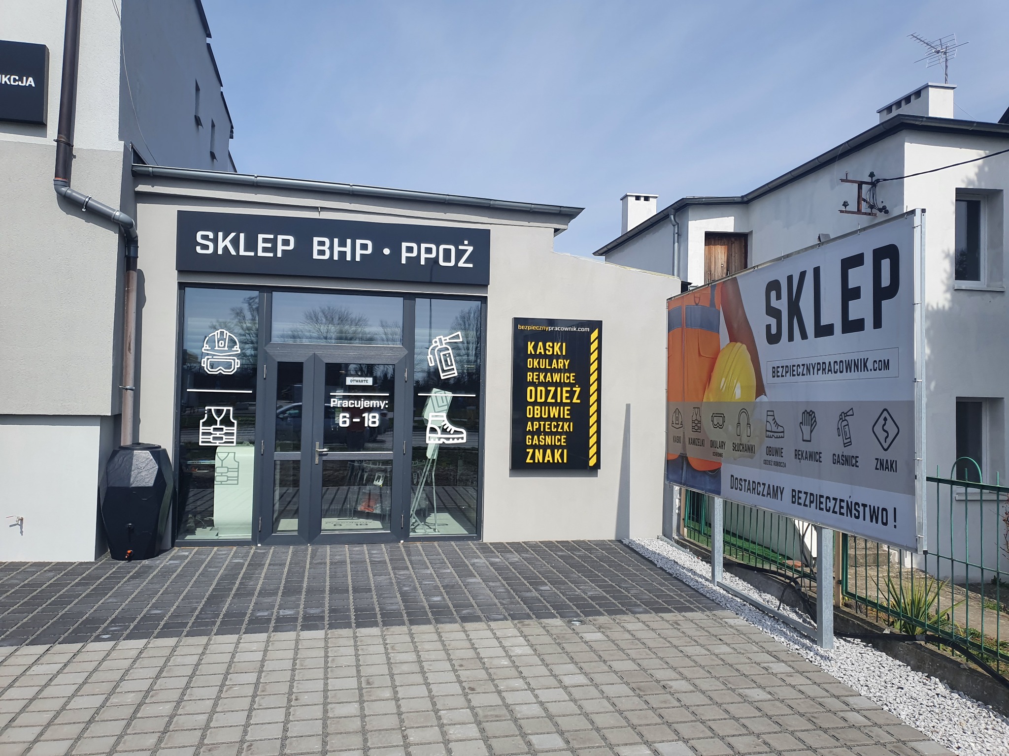 Fasada sklepu BHP i PPOŻ z dużymi witrynami, prezentująca odzież i akcesoria ochronne, tablica informacyjna z godzinami otwarcia i logo firmy oraz baner reklamowy z hasłem 'Dostarczamy...
