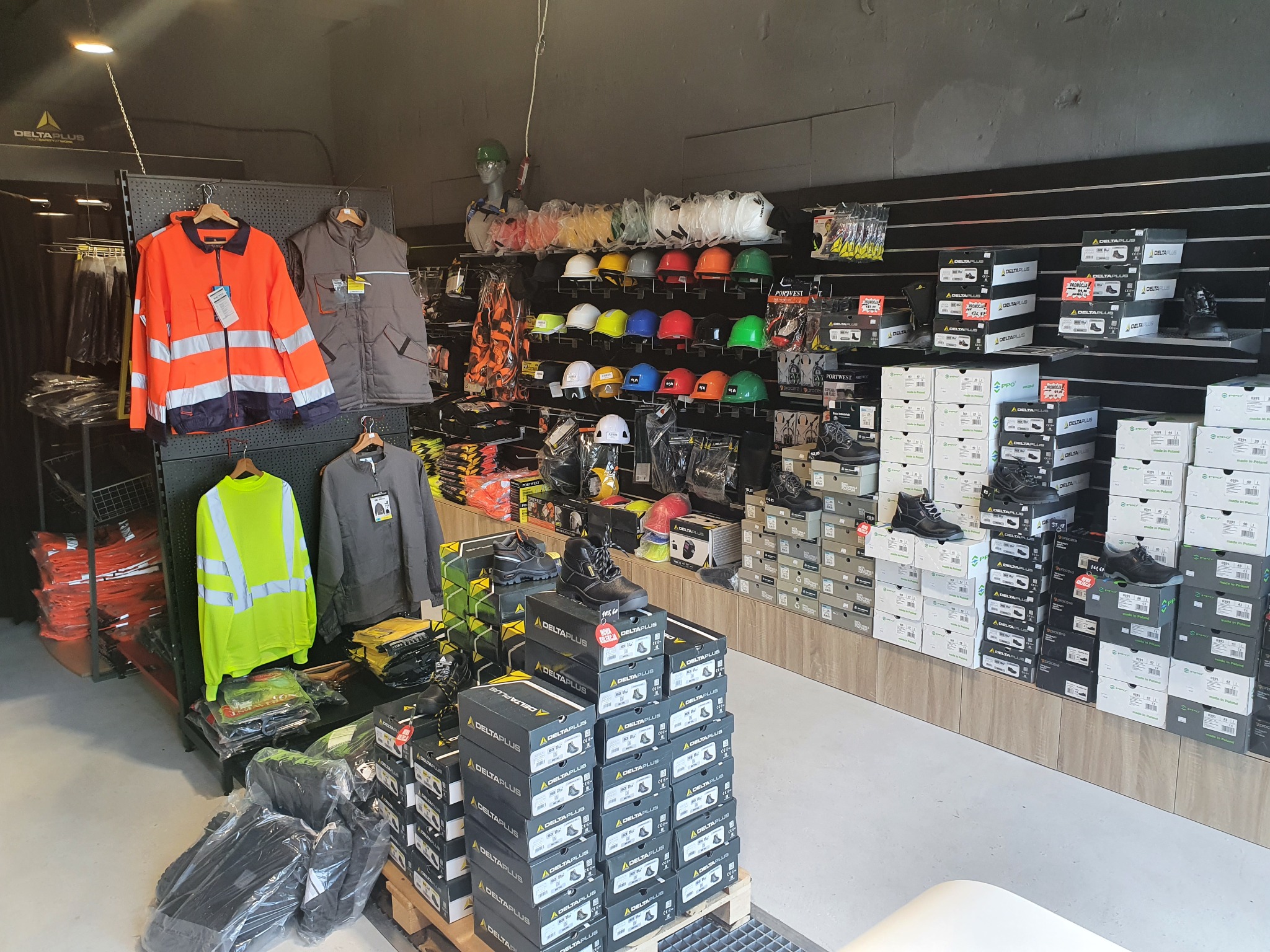 Wnętrze sklepu z odzieżą i obuwiem roboczym: kurtki ostrzegawcze w kolorze pomarańczowym i żółtym, kamizelki, kaski ochronne w różnych kolorach, buty robocze w pudełkach Delta Plus ułożone...