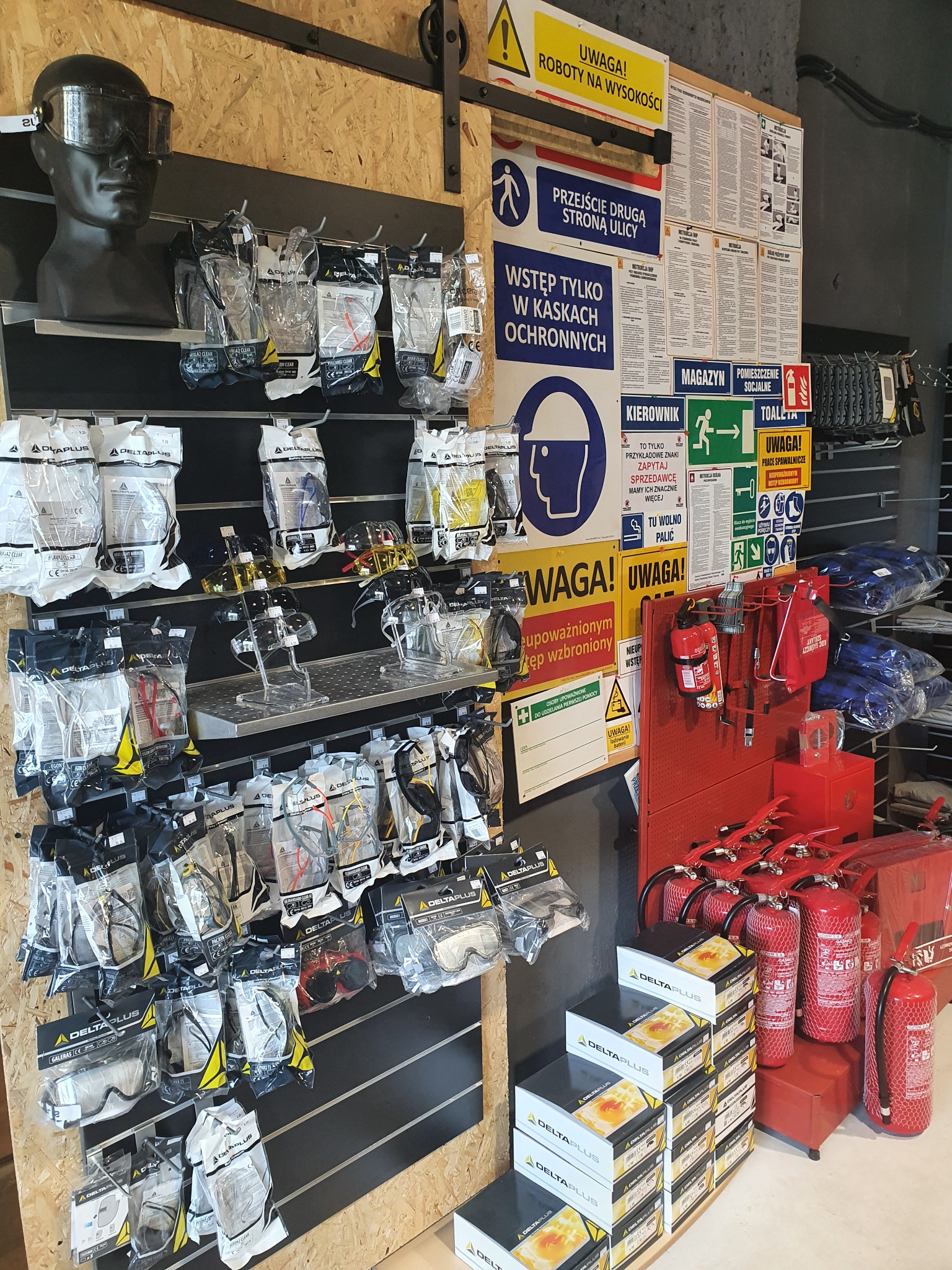 Ekspozycja środków ochrony osobistej: okulary ochronne, maski, rękawice i gaśnice Delta Plus, wraz z tablicami informacyjnymi BHP i znakami ewakuacyjnymi.