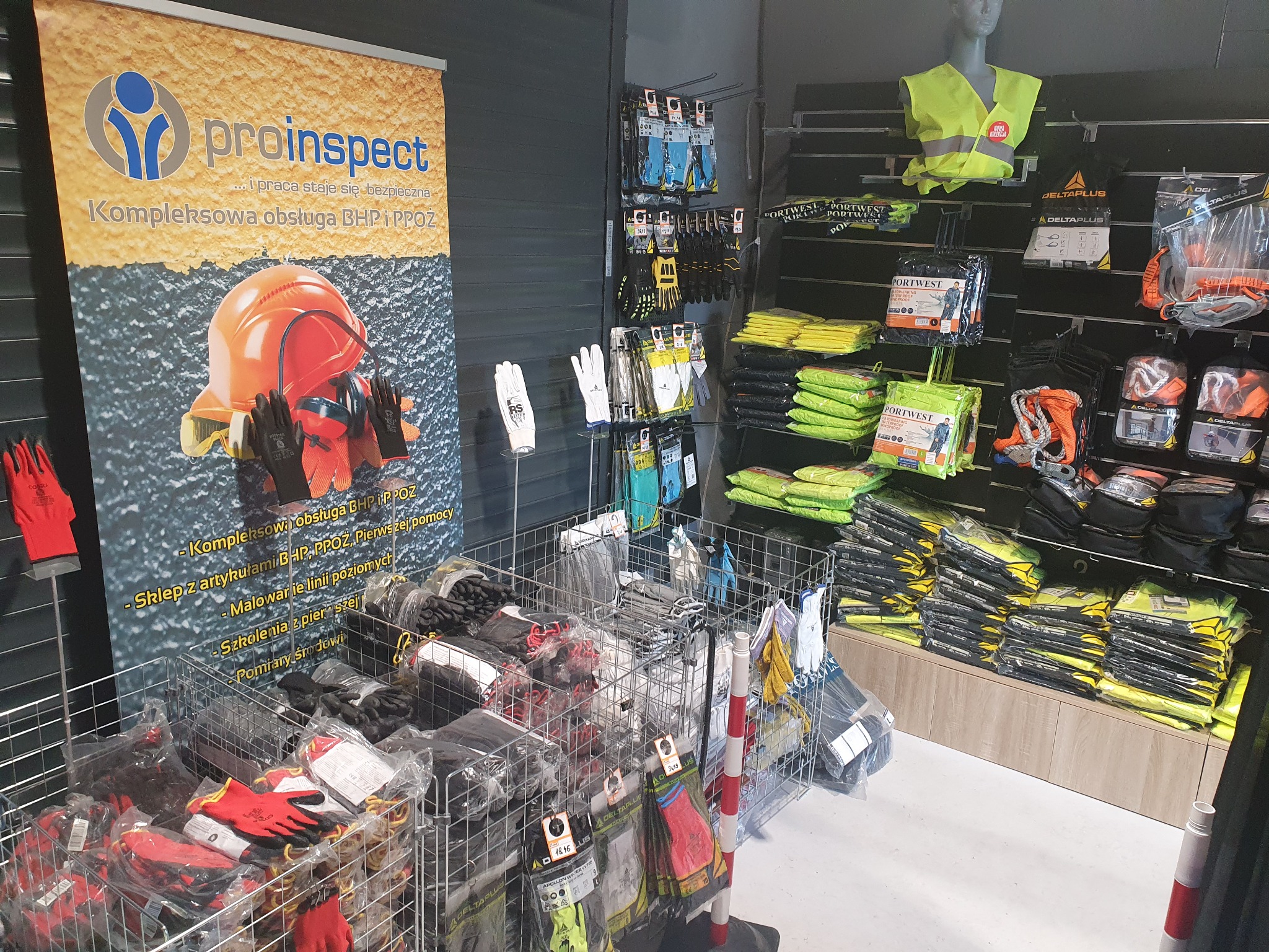 Ekspozycja sklepu z artykułami BHP i PPOŻ: Rękawice robocze w koszach, kamizelki odblaskowe na manekinie i półkach, baner reklamowy firmy Proinspect z hełmem i ochronnikami słuchu.