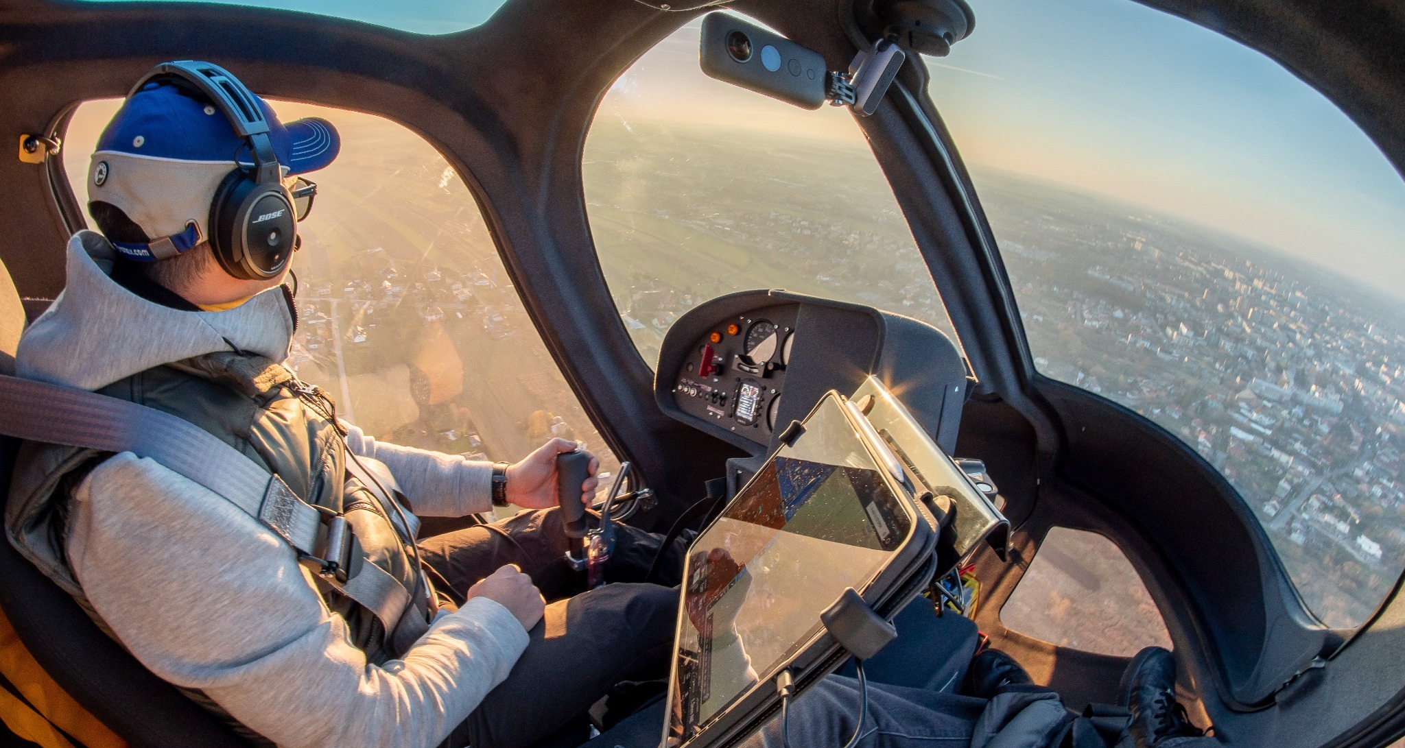 Pilot w kokpicie helikoptera podczas lotu nad miastem, widok zza pleców, w tle zabudowa miejska i zachodzące słońce.