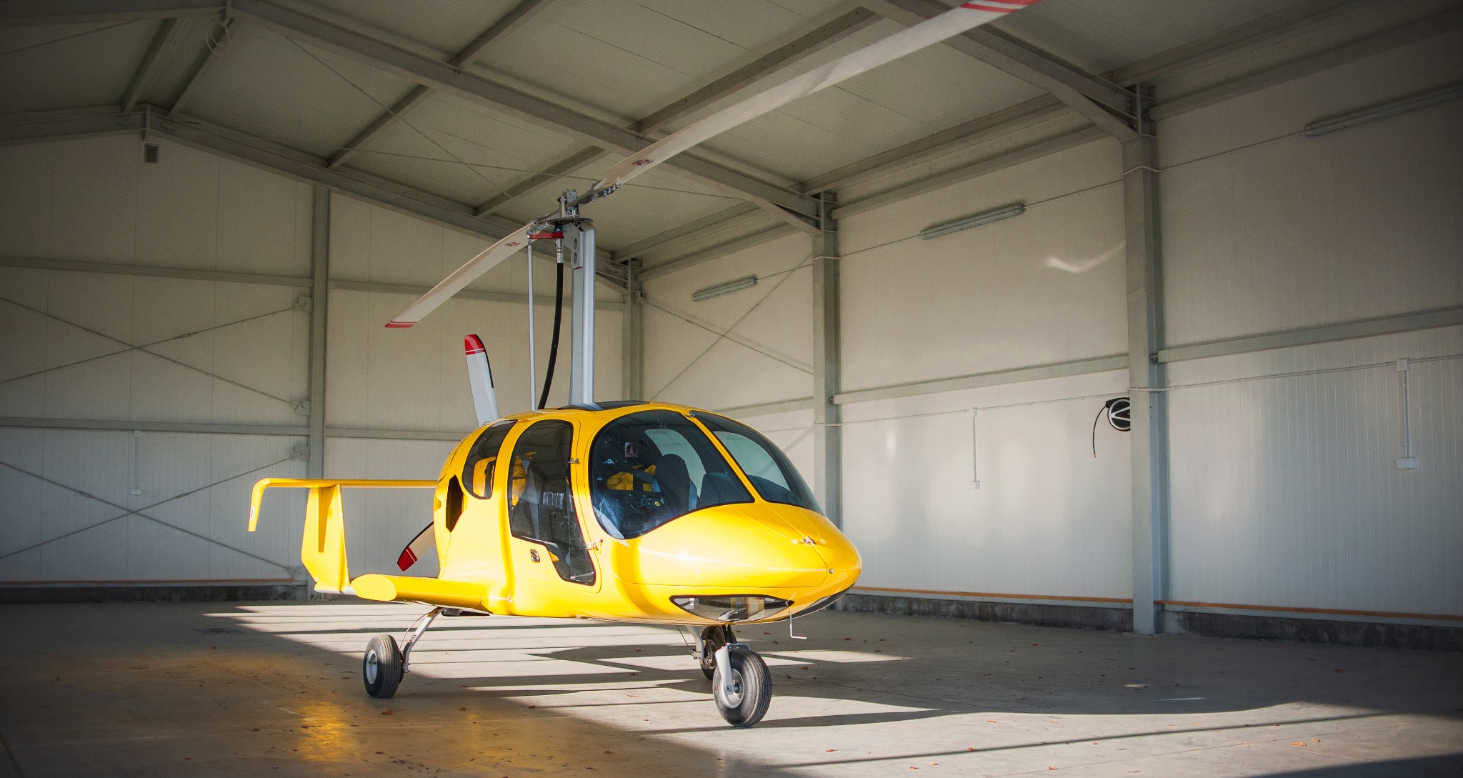 Żółty wiatrakowiec zaparkowany wewnątrz hangaru z metalowymi ścianami i betonową podłogą, widoczne ślady opon.
