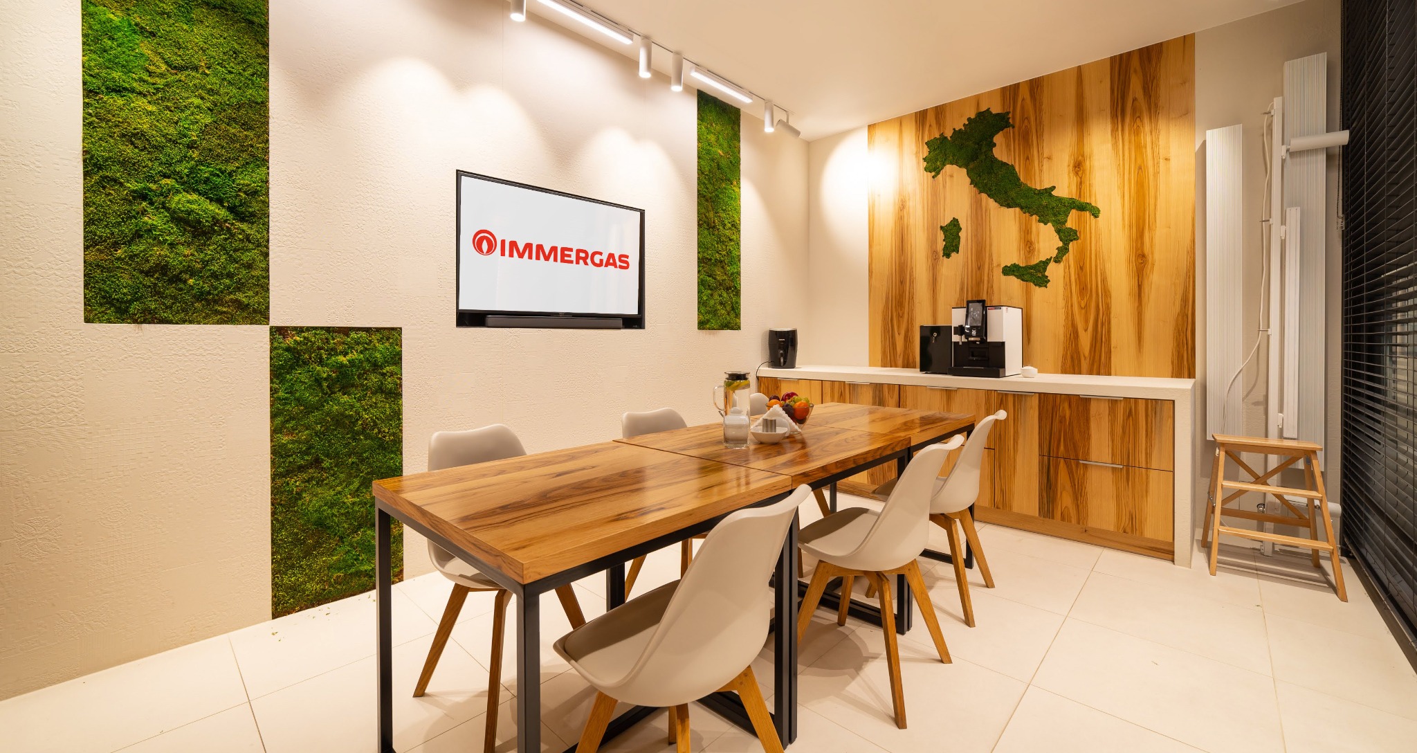 Nowoczesna sala konferencyjna z drewnianym stołem, białymi krzesłami, ścianą z mchu i drewnianą mapą Włoch. Na ścianie wisi telewizor z logo firmy Immergas.