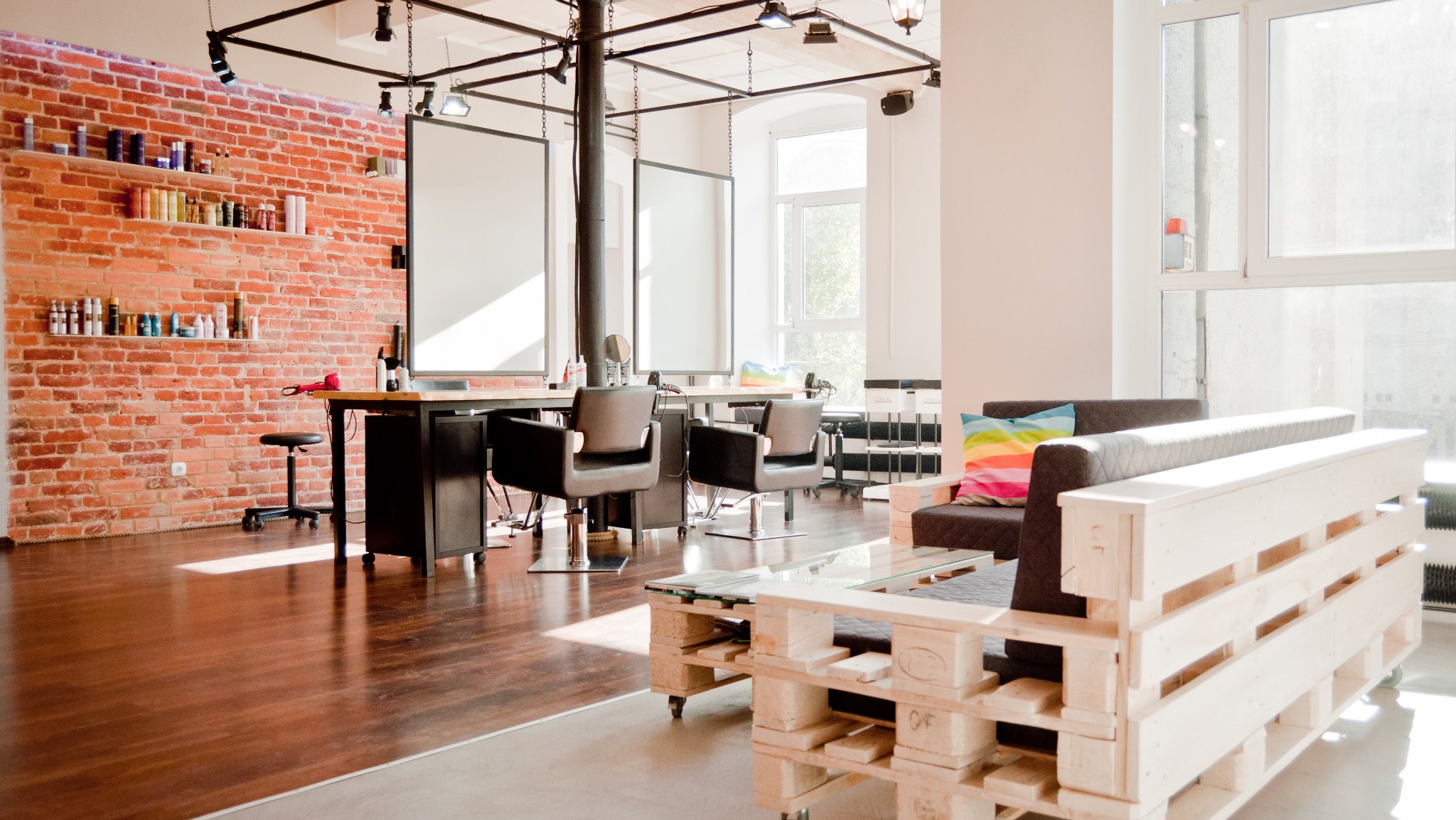 Jasne wnętrze salonu fryzjerskiego z ceglaną ścianą, drewnianą podłogą, stanowiskami pracy z lustrami i krzesłami, oraz strefą oczekiwania z sofą z palet i kolorową poduszką.