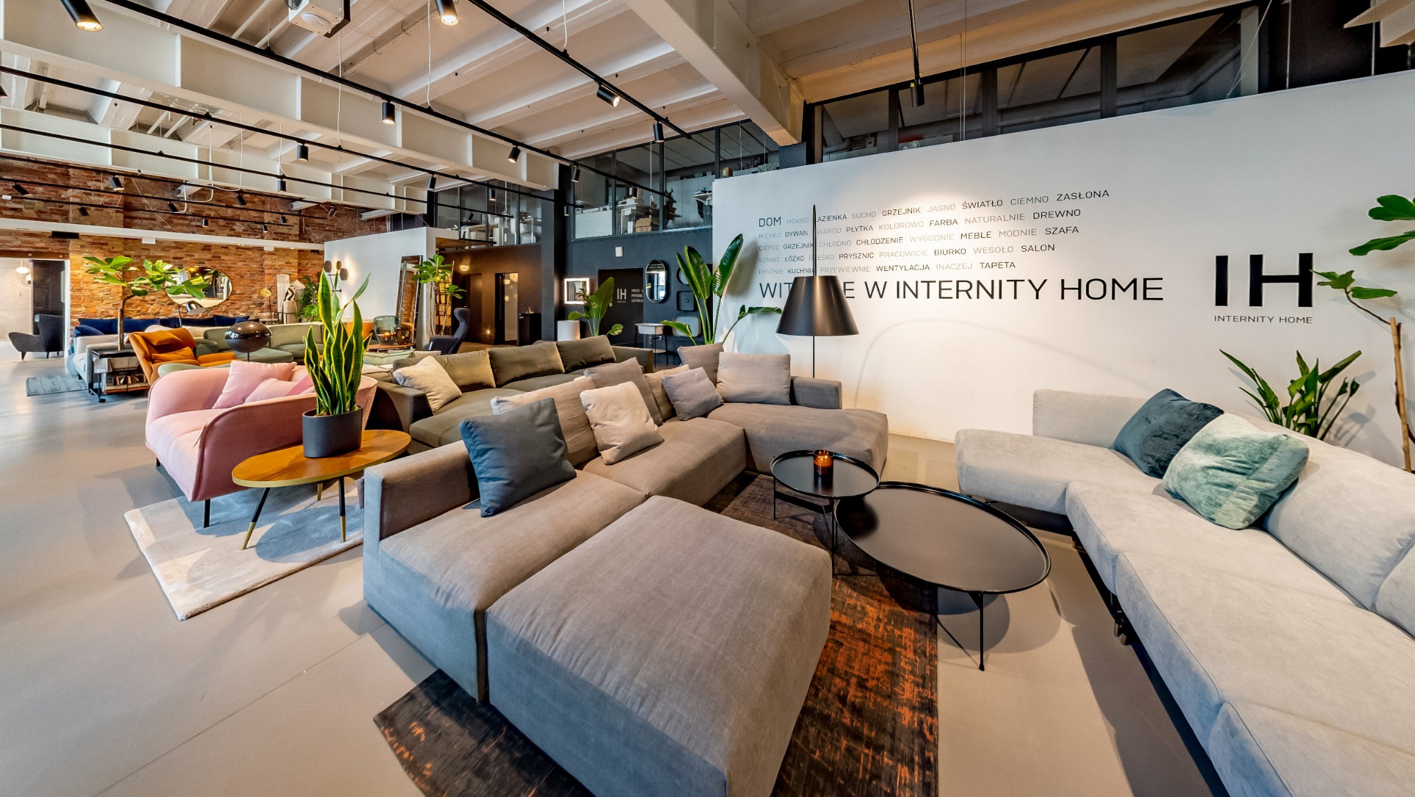 Wnętrze sklepu meblowego z ekspozycją różnorodnych sof, foteli i stolików kawowych; na ścianie logo 'Internity Home' i wypisane słowa kluczowe związane z wyposażeniem wnętrz.