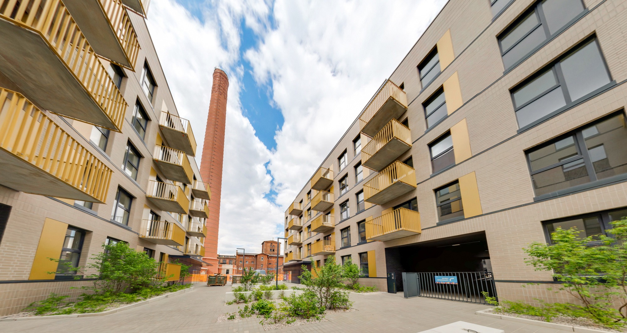 Dziedziniec nowoczesnego kompleksu mieszkalnego z ceglanymi budynkami, żółtymi balkonami i wysokim, ceglanym kominem w tle, widziany z poziomu chodnika.