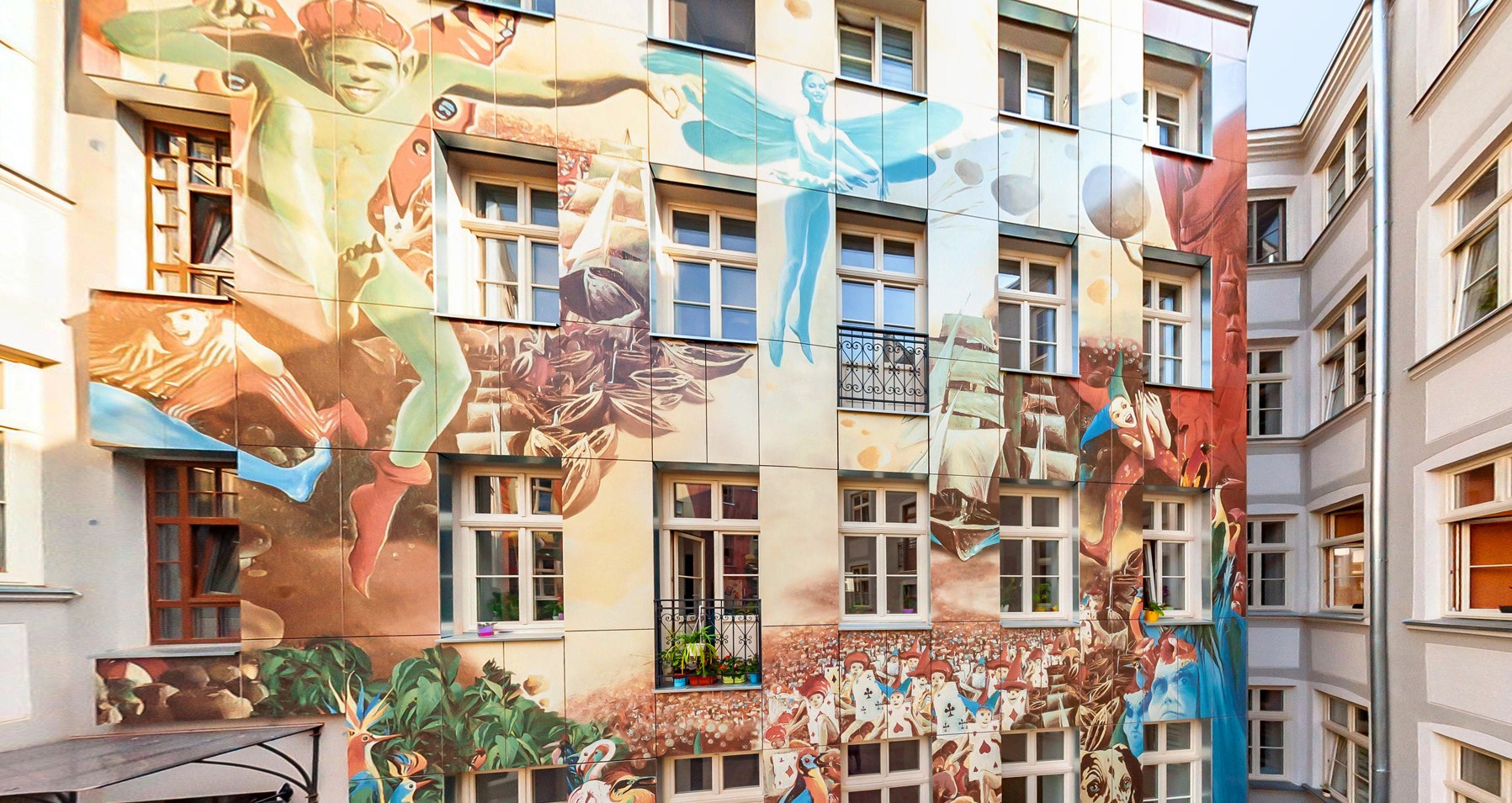 Fasada budynku pokryta wielobarwnym muralem z motywami postaci, żaglowców i kart do gry, widoczna z wewnętrznego dziedzińca.