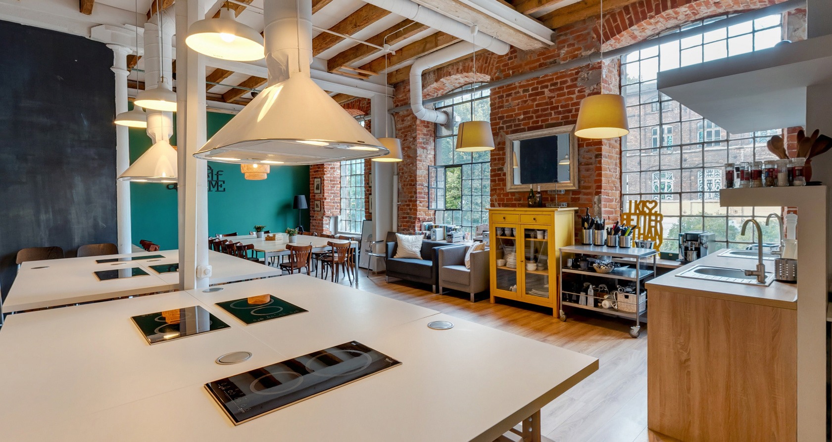 Przestronne wnętrze loftu z długimi stołami z wbudowanymi płytami grzewczymi, kuchnią z oknem w ceglanej ścianie, szarą sofą i żółtą szafką.