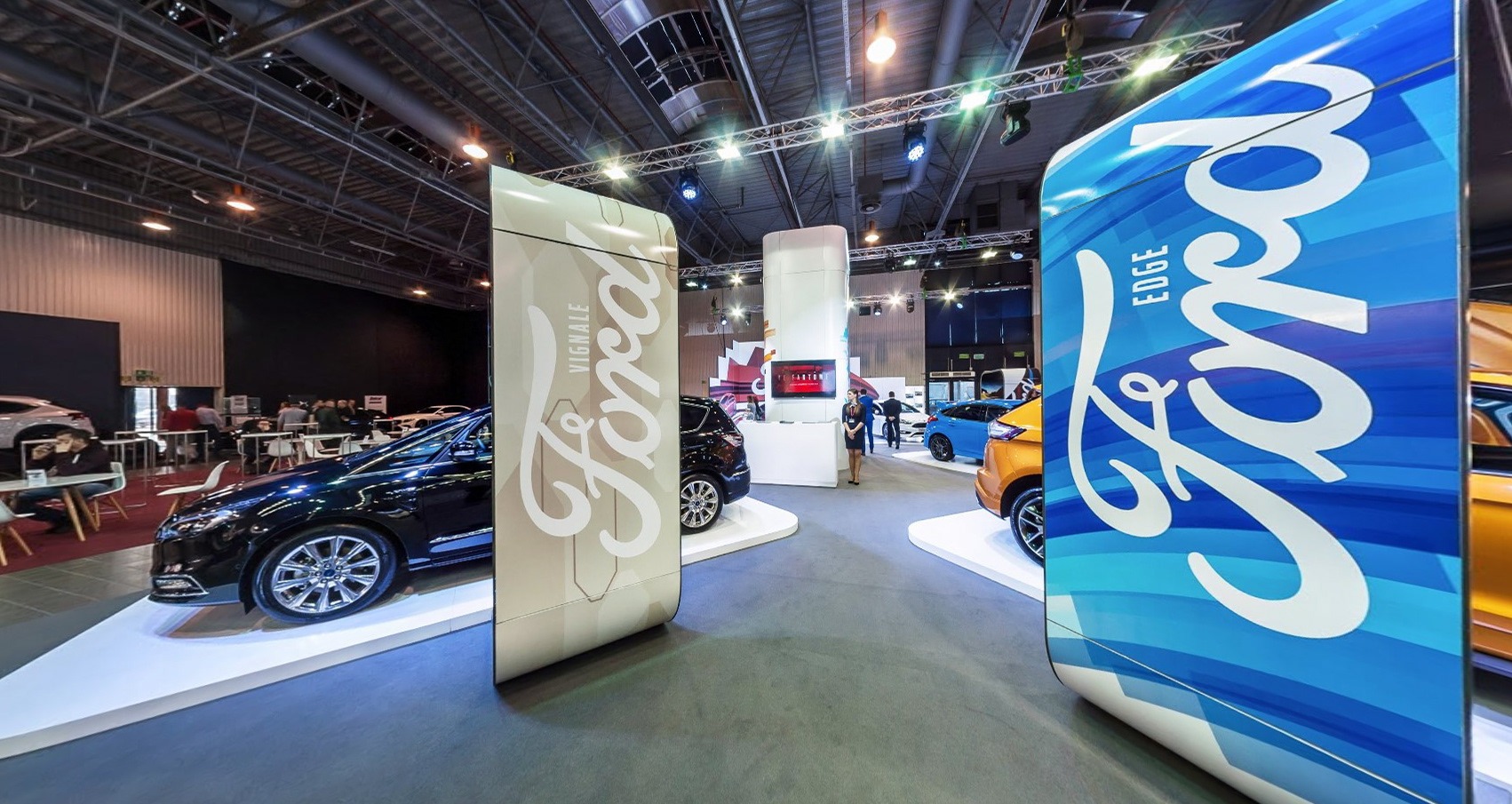 Wnętrze hali wystawienniczej z samochodami Ford na podestach, logo firmy na panelach i osoby w tle.