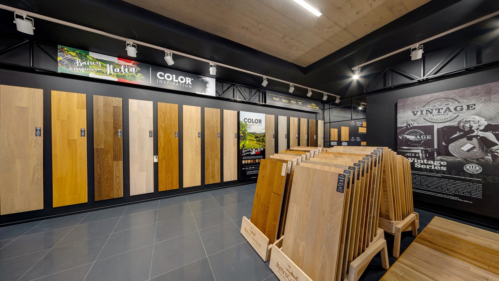 Ekspozycja paneli podłogowych w sklepie: różnorodność kolorów i wzorów drewna, oświetlenie punktowe, plakaty reklamowe na ścianach.