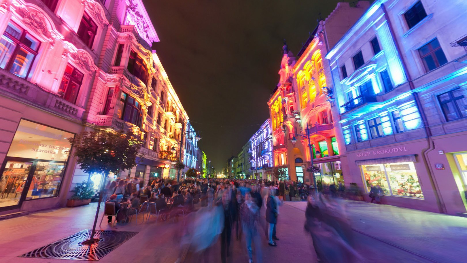 Ulica w nocy z budynkami oświetlonymi kolorowymi światłami, widoczne rozmyte sylwetki ludzi.
