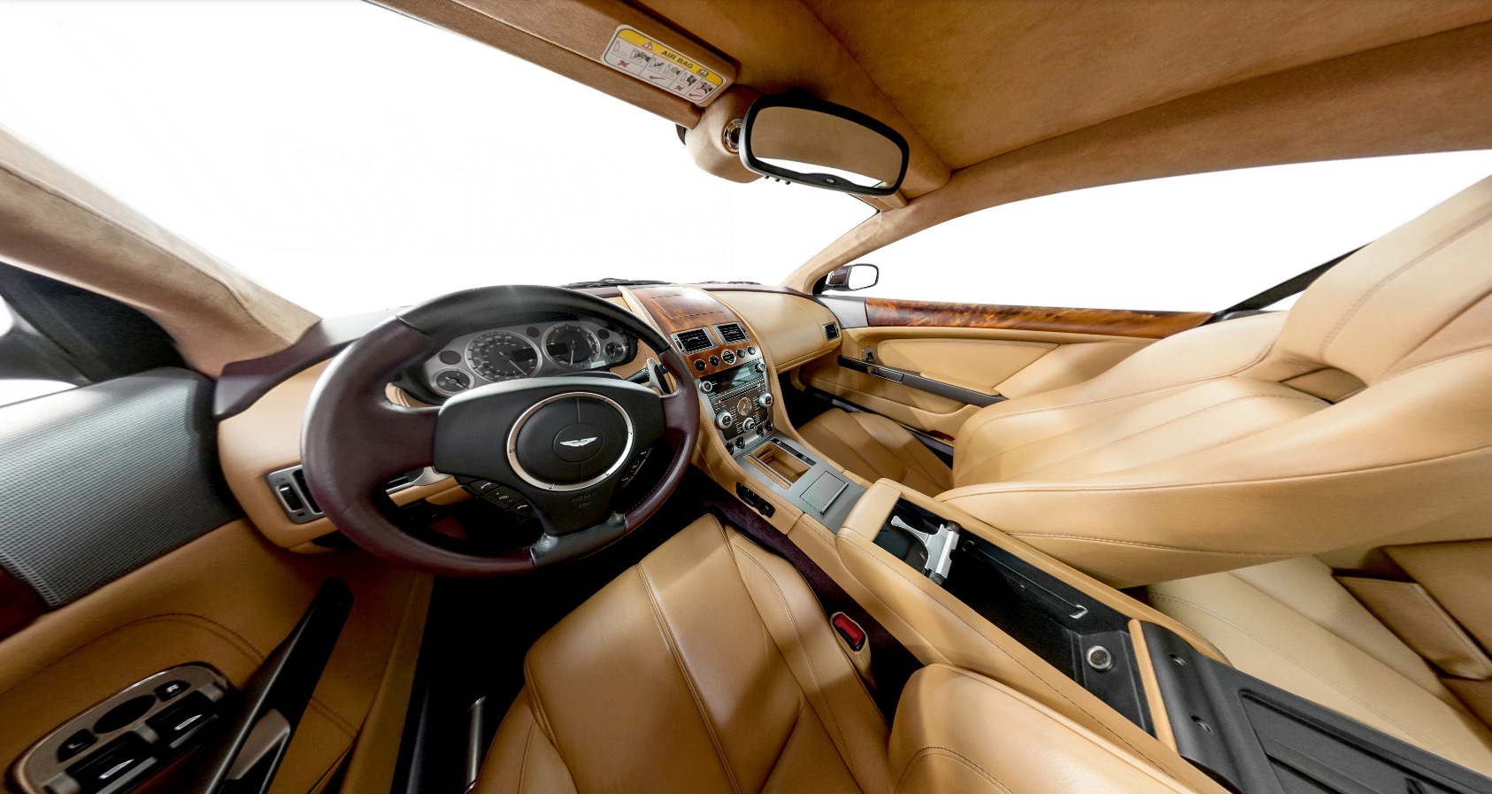 Wnętrze luksusowego samochodu sportowego z beżową skórzaną tapicerką, drewnianymi elementami wykończenia deski rozdzielczej, czarną kierownicą z chromowanym logo producenta oraz widocznym...