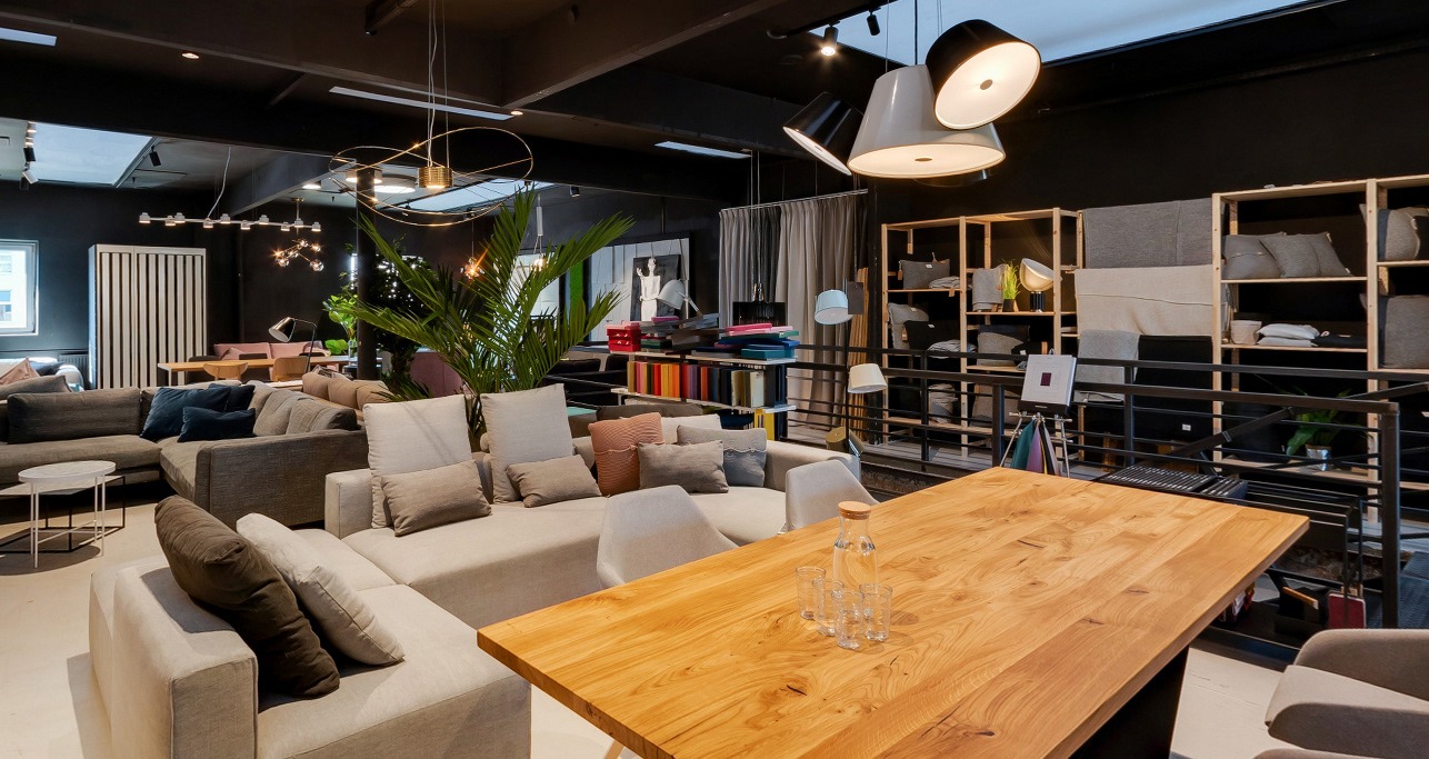 Jasne wnętrze salonu z narożną sofą, drewnianym stołem i dekoracyjnymi lampami wiszącymi; widoczne regały z tekstyliami i roślina doniczkowa.