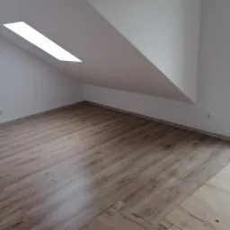 Poddasze z oknem dachowym i nowo położonymi panelami podłogowymi w odcieniach brązu, zabezpieczonymi częściowo kartonem.