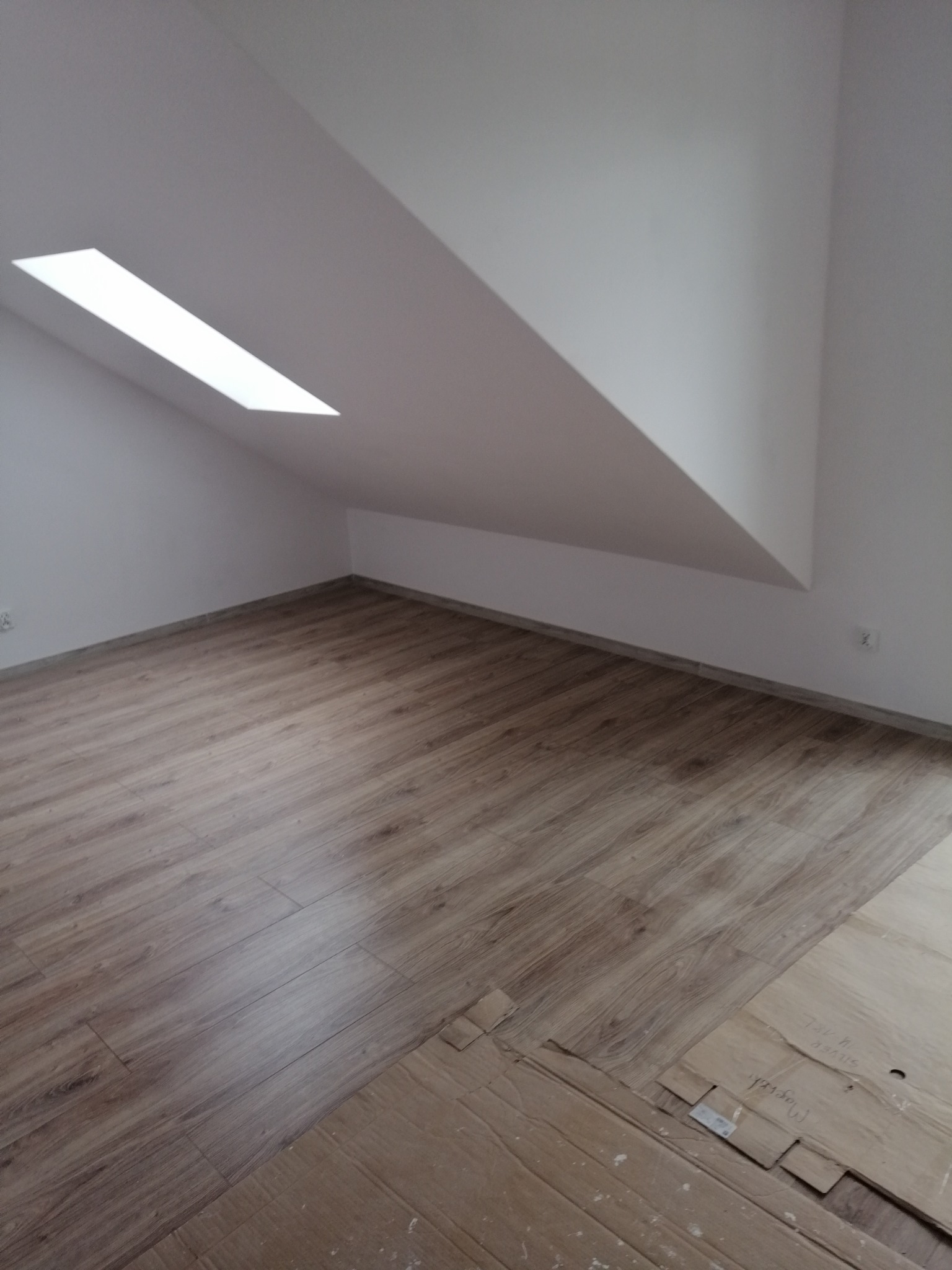 Poddasze z oknem dachowym i nowo położonymi panelami podłogowymi w odcieniach brązu, zabezpieczonymi częściowo kartonem.