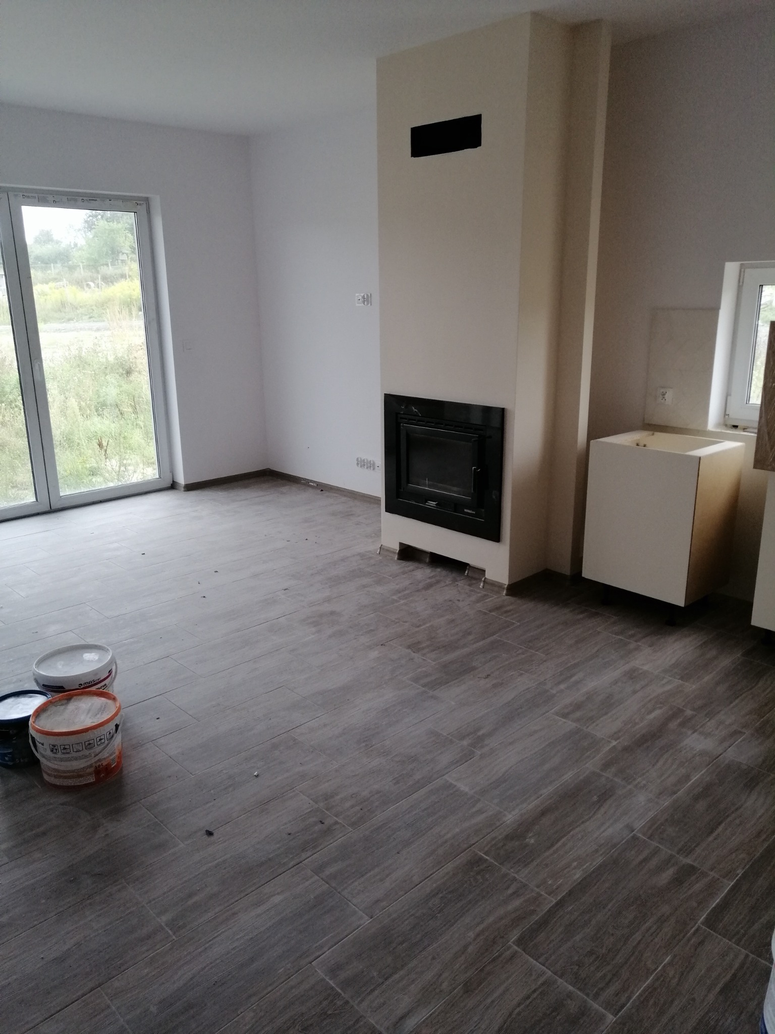 Wnętrze w trakcie wykończenia: ułożona podłoga z płytek imitujących drewno, w tle kominek z czarną obudową i niedokończona zabudowa kuchenna, widoczne puszki z farbą.