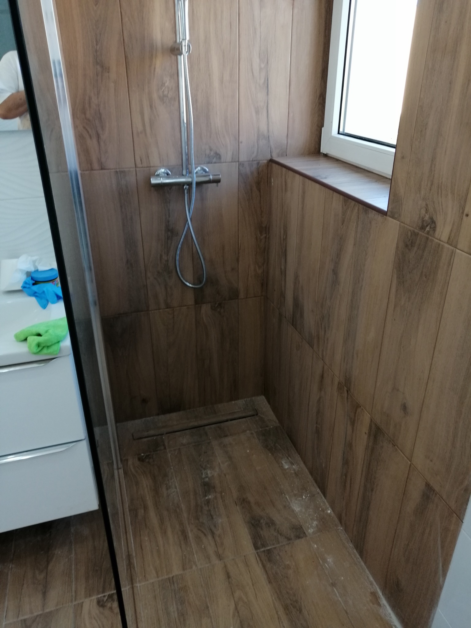 Kabina prysznicowa wyłożona płytkami imitującymi drewno, z widoczną armaturą i oknem, częściowo widoczna umywalka z akcesoriami.