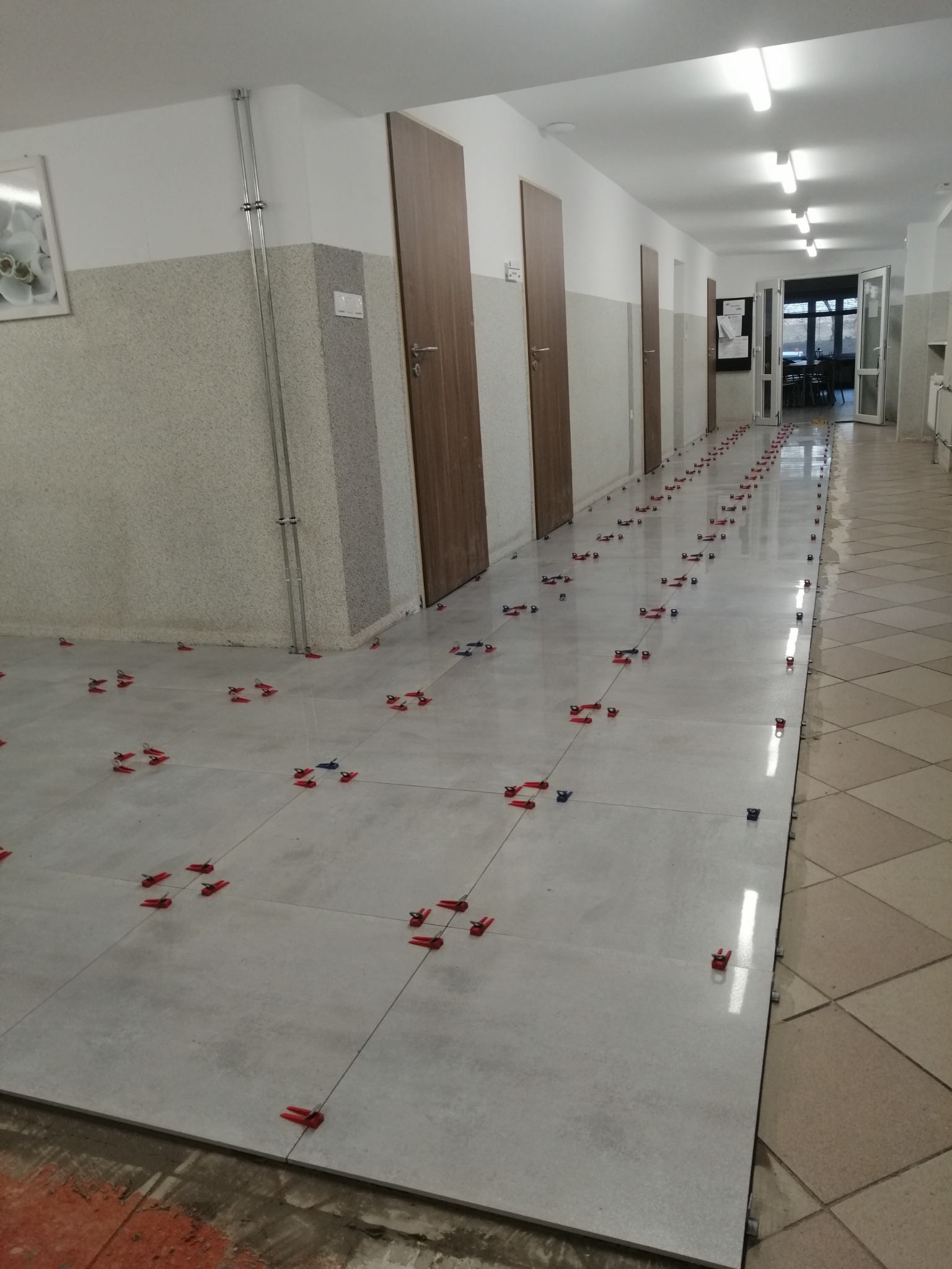 Korytarz w trakcie układania jasnoszarych płytek podłogowych z użyciem czerwonych i niebieskich klipsów poziomujących, widoczne drzwi i oświetlenie sufitowe.