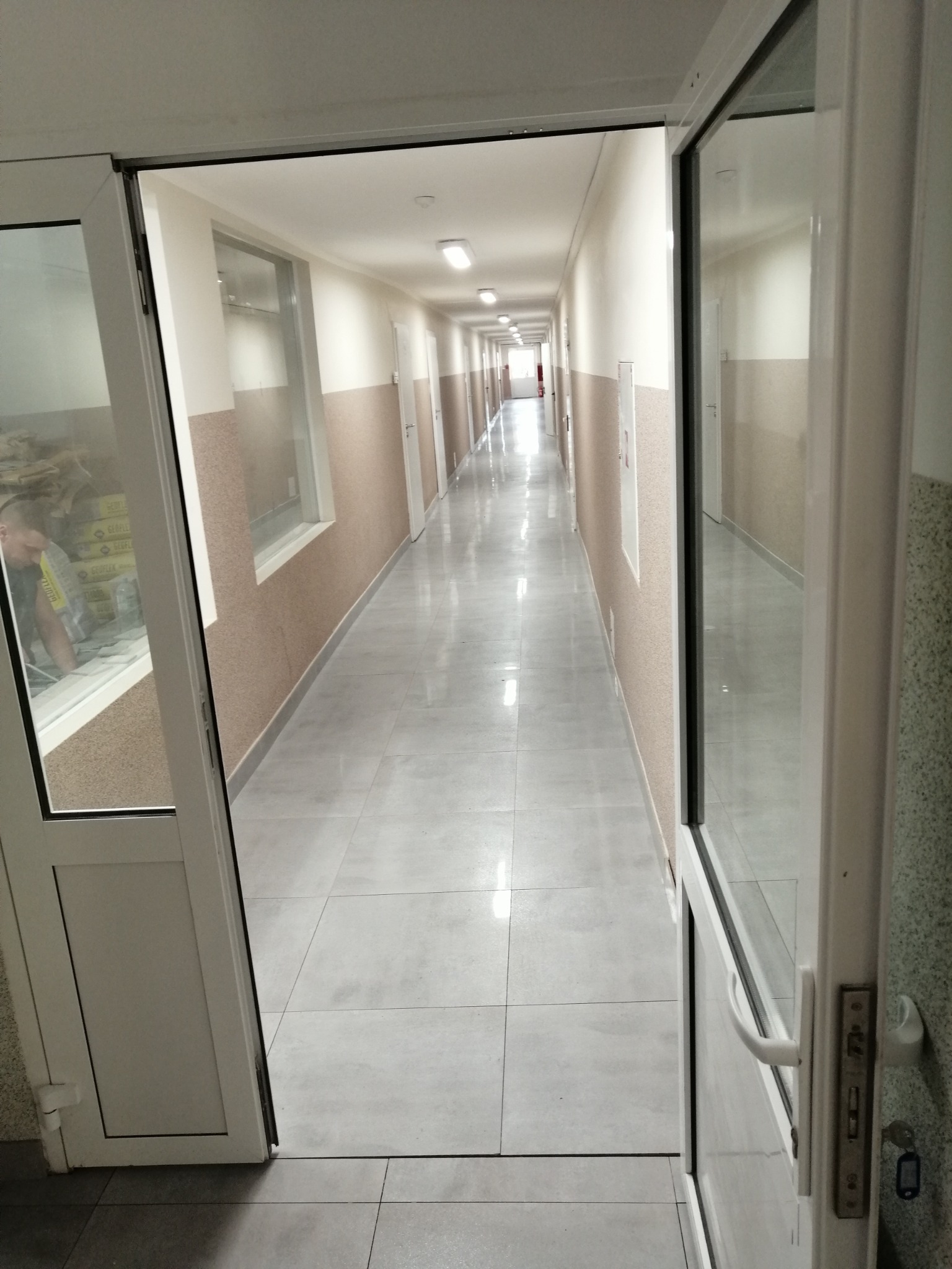 Długi korytarz w budynku w Starogardzie Gdańskim, wyłożony szarymi płytkami podłogowymi, widoczny przez otwarte białe drzwi. W tle, w oknie, widoczny pracownik.