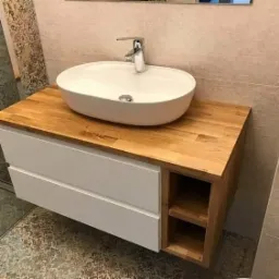 Biała, owalna umywalka nablatowa z chromowaną baterią na blacie z jasnego drewna, podwieszana szafka z białymi szufladami i otwartymi półkami.