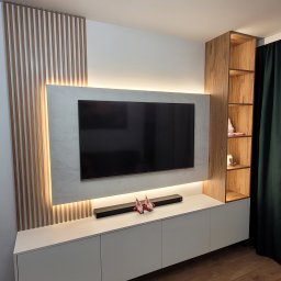 Stylove Kuchnie - Nowoczesna ściana telewizyjna z białymi szafkami, podświetlanym panelem z efektem marmuru, drewnianymi lamelami i otwartym regałem z forniru w kolorze drewna.