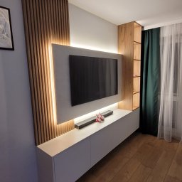 Stylove Kuchnie - Nowoczesna zabudowa ściany telewizyjnej z drewnianymi lamelami, podświetleniem LED, białą szafką RTV i półkami z jasnego drewna.