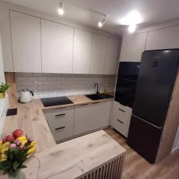 Jasna kuchnia z drewnianym blatem, czarnym zlewem i sprzętem AGD, oświetlona punktowymi lampami sufitowymi, z owocami i kwiatami na blacie.