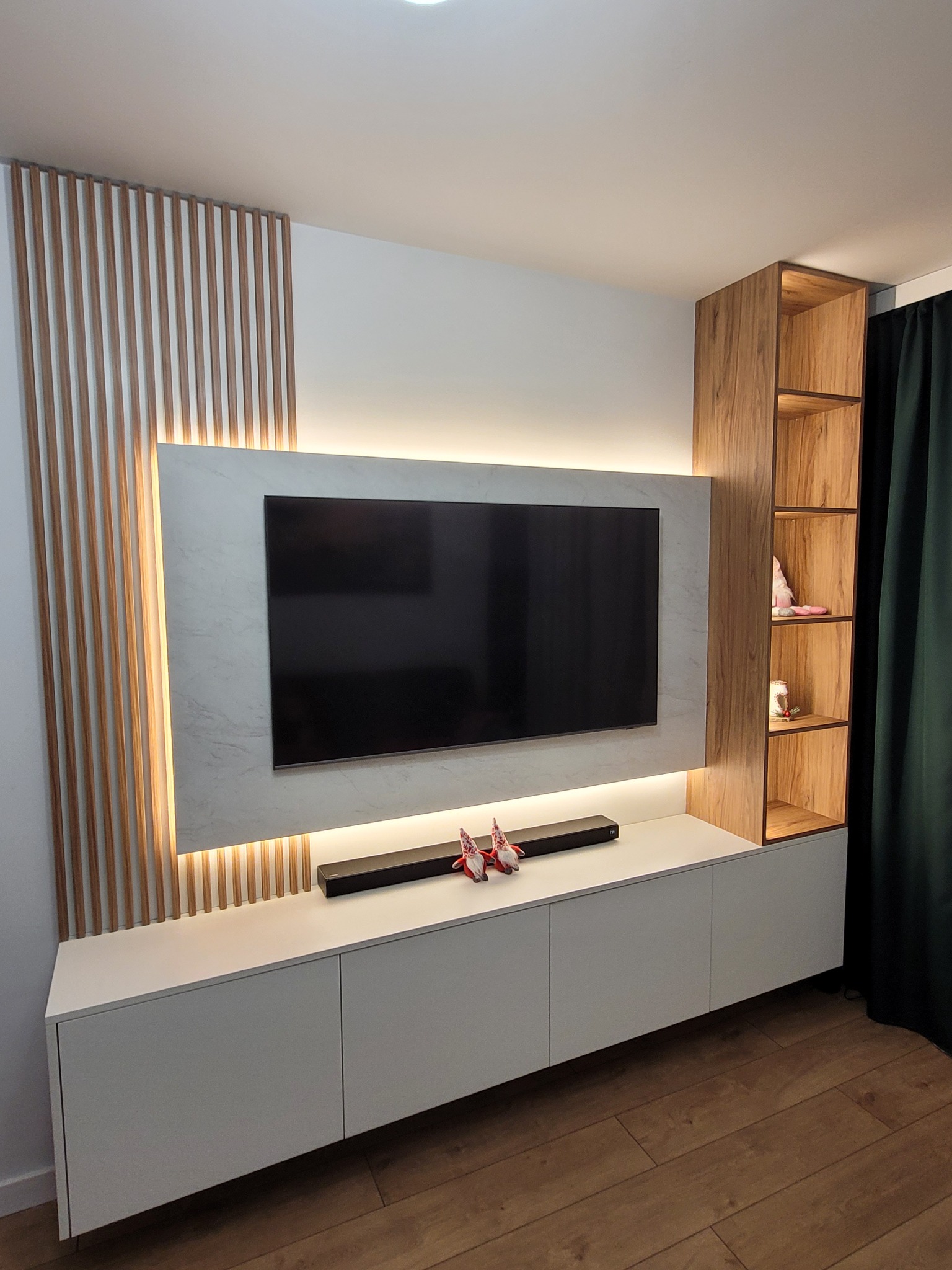 Nowoczesna ściana telewizyjna z białymi szafkami, podświetlanym panelem z efektem marmuru, drewnianymi lamelami i otwartym regałem z forniru w kolorze drewna.