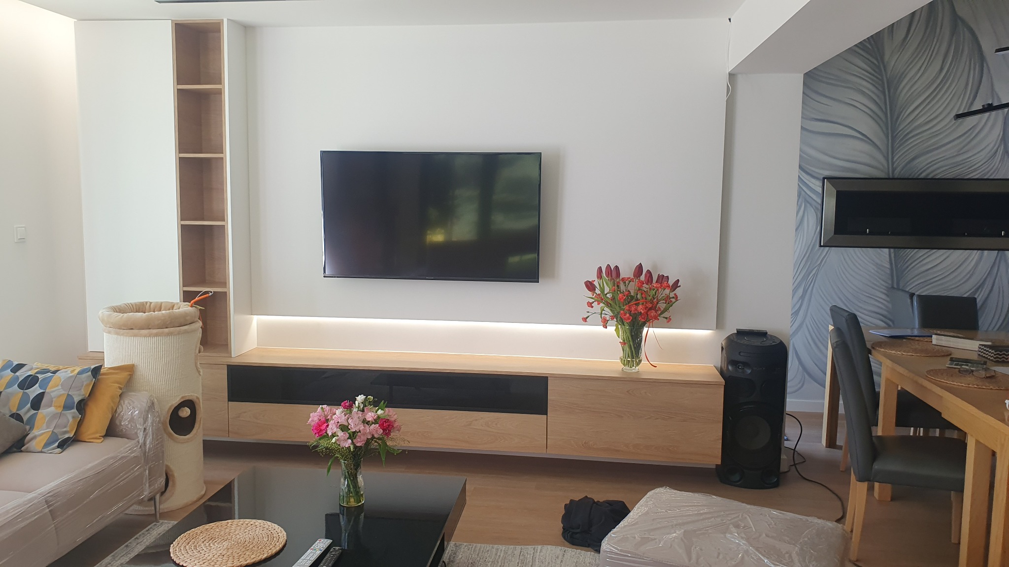 Nowoczesny salon z zabudową meblową: białe szafy, szafka RTV z podświetleniem LED, telewizor na ścianie, dekoracyjny kominek elektryczny, kwiaty w wazonie. Widoczny stolik kawowy i fragment sofy...