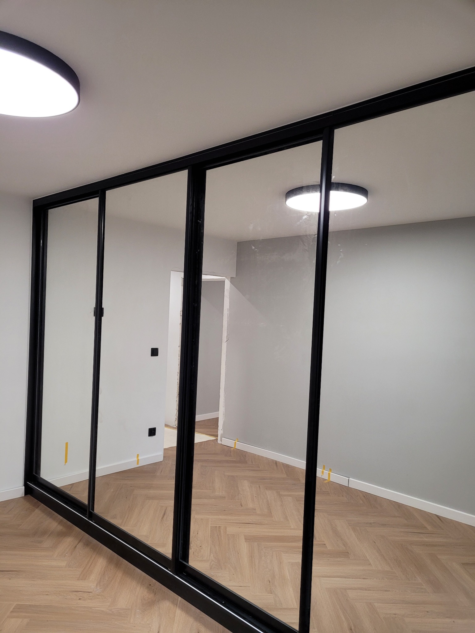 Czarna, przesuwna szafa z lustrami wnękowa na tle jasnego parkietu i szarej ściany, widoczne taśmy malarskie zabezpieczające listwy.