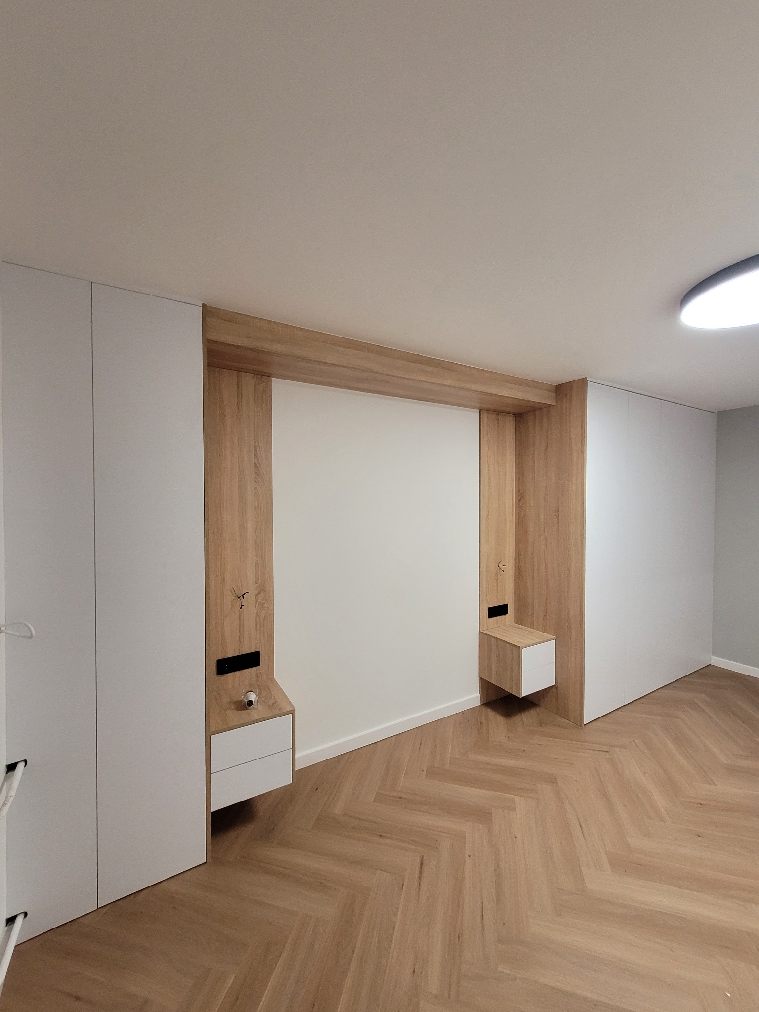 Nowoczesna sypialnia z zabudową meblową: białe szafy wnękowe po bokach, drewniana rama z szafkami nocnymi, parkiet w jodełkę.