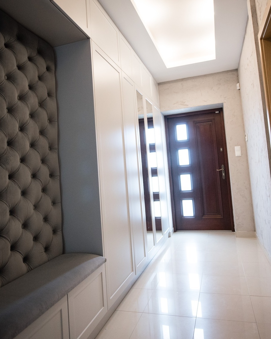 Biała szafa wnękowa z lustrami i tapicerowanym siedziskiem w przedpokoju, widok z perspektywy podłogi.