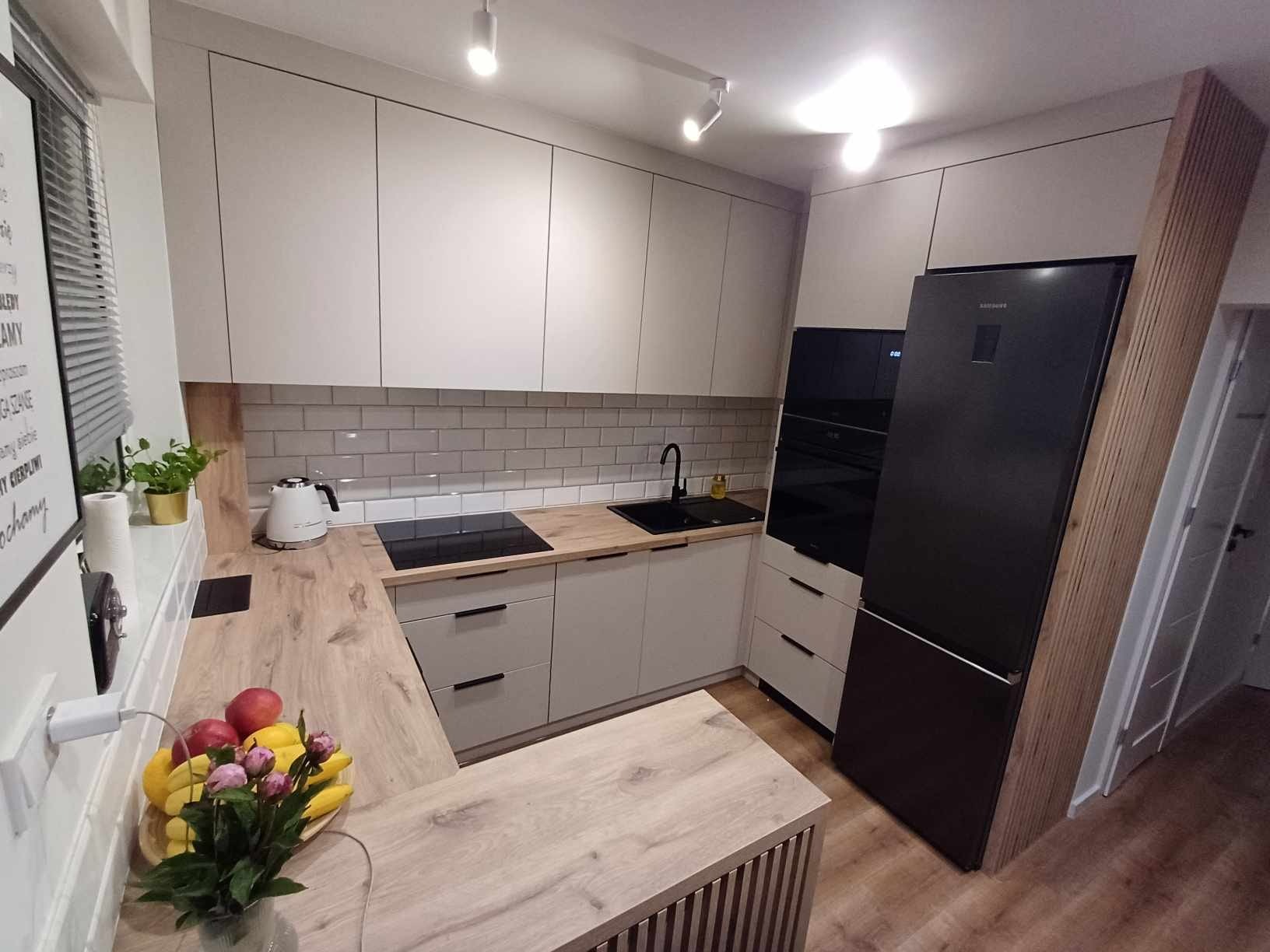 Jasna kuchnia z drewnianym blatem, czarnym zlewem i sprzętem AGD, oświetlona punktowymi lampami sufitowymi, z owocami i kwiatami na blacie.