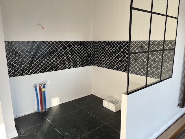 Łazienka w trakcie wykańczania: czarno-białe płytki wzorzyste na ścianie, czarna podłoga, widoczne rury instalacyjne i szklana ścianka działowa w stylu industrialnym.