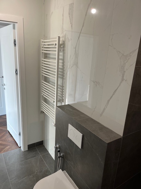 Wykończona łazienka z białymi płytkami imitującymi marmur, szarymi płytkami podłogowymi i zabudową toalety, grzejnikiem drabinkowym oraz fragmentem białej ściany i drewnianej podłogi w tle.