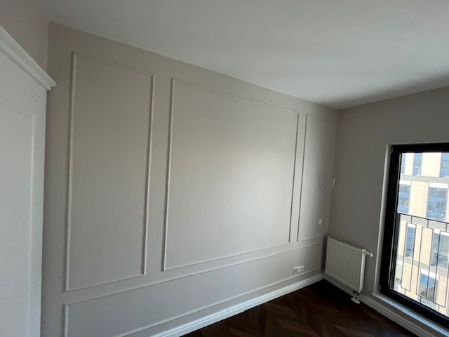 Beżowa ściana z dekoracyjnymi listwami w klasycznym stylu, widoczny fragment podłogi z parkietem w jodełkę, grzejnik i okno z widokiem na budynek.