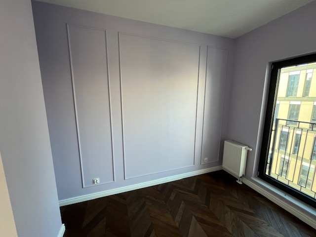 Pokój po malowaniu z dekoracyjnymi listwami ściennymi w odcieniach fioletu, parkiet w jodełkę i grzejnik pod oknem z widokiem na budynek.