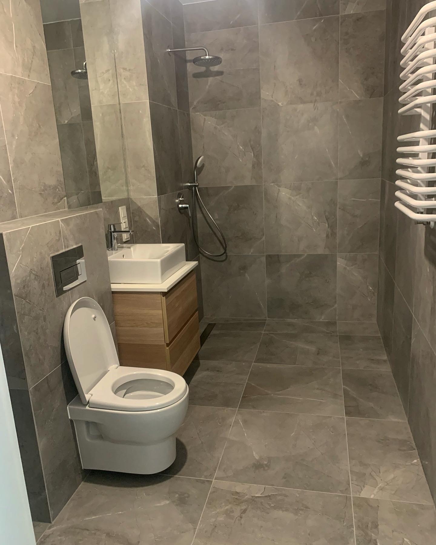 Nowoczesna łazienka w odcieniach szarości z kabiną prysznicową typu walk-in, wiszącą toaletą i umywalką nablatową na drewnianej szafce, wykończona płytkami imitującymi marmur.