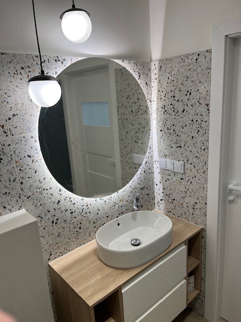 Nowoczesna łazienka z okrągłym lustrem podświetlanym LED, umywalką nablatową na szafce z drewnianym blatem i białymi frontami oraz ścianami wyłożonymi płytkami imitującymi lastryko.