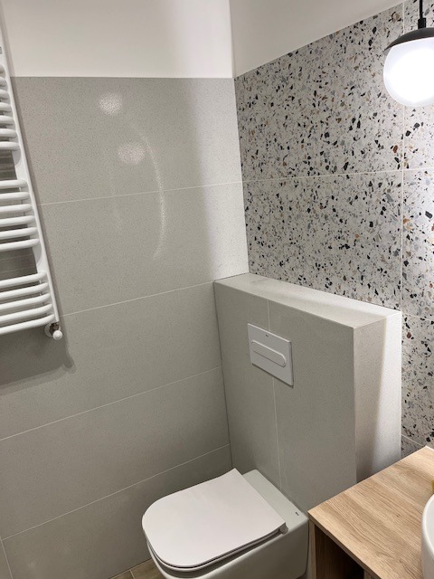 Nowoczesna łazienka z szarymi płytkami imitującymi kamień, toaleta podwieszana z zabudową, grzejnik drabinkowy i ściana z mozaiką lastryko.