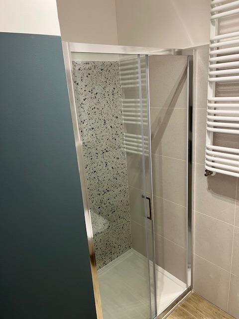 Nowoczesna kabina prysznicowa z przesuwnymi drzwiami, wykończona płytkami w mozaikę, z widocznym grzejnikiem drabinkowym i fragmentem ściany pomalowanej na kolor morski.