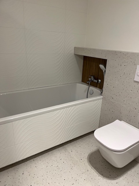 Nowoczesna łazienka z wanną obudowaną płytkami z falistym wzorem, toaletą podwieszaną i ścianą wykończoną płytkami imitującymi lastryko. Wnęka nad wanną wyłożona drewnem.