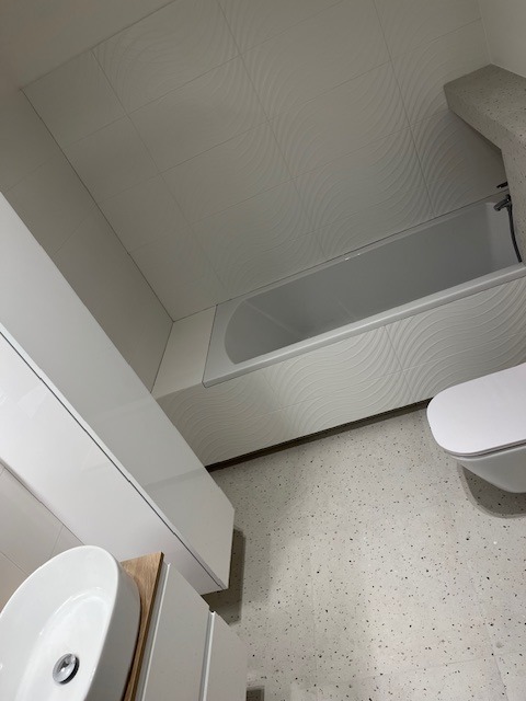 Biała łazienka po remoncie w Warszawie: widok z góry na wannę obudowaną płytkami z delikatnym wzorem fali, umywalkę nablatową na szafce z drewnianym blatem i toaletę, całość utrzymana w jasnej...