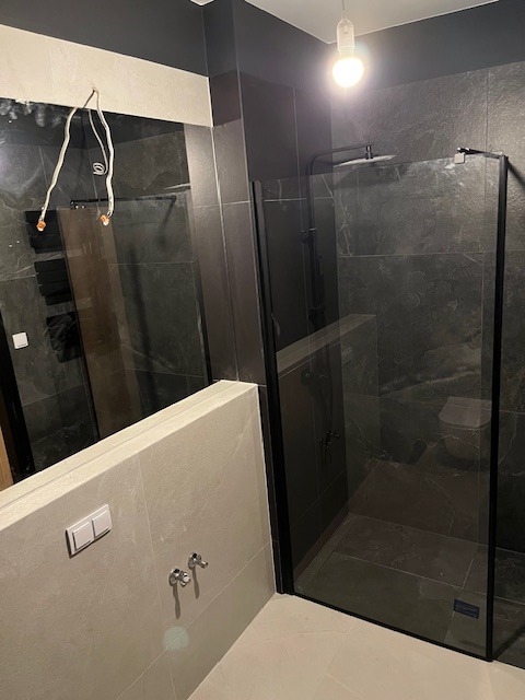 Nowoczesna łazienka z kabiną prysznicową typu walk-in z czarnymi profilami, szare płytki imitujące kamień, podtynkowa bateria umywalkowa i wiszące WC widoczne przez szybę.