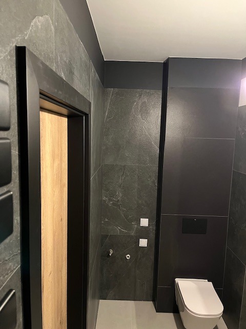 Nowoczesna łazienka w odcieniach szarości i czerni z drewnianymi akcentami w drzwiach, minimalistyczna toaleta podwieszana, marmurowe płytki i czarne listwy wykończeniowe.