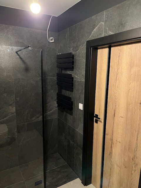Nowoczesna łazienka z ciemnoszarymi płytkami imitującymi kamień, kabiną prysznicową typu walk-in z czarnymi profilami, czarnym grzejnikiem drabinkowym i drewnianymi drzwiami z czarną ościeżnicą.