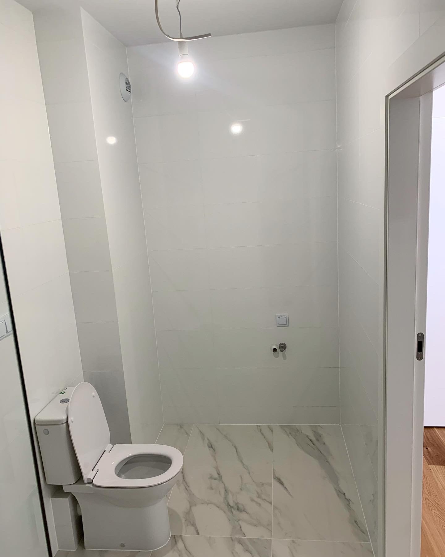 Nowoczesna, biała łazienka z marmurową podłogą i wiszącą lampą halogenową, prezentująca świeżo wykończone wnętrze z widoczną toaletą.