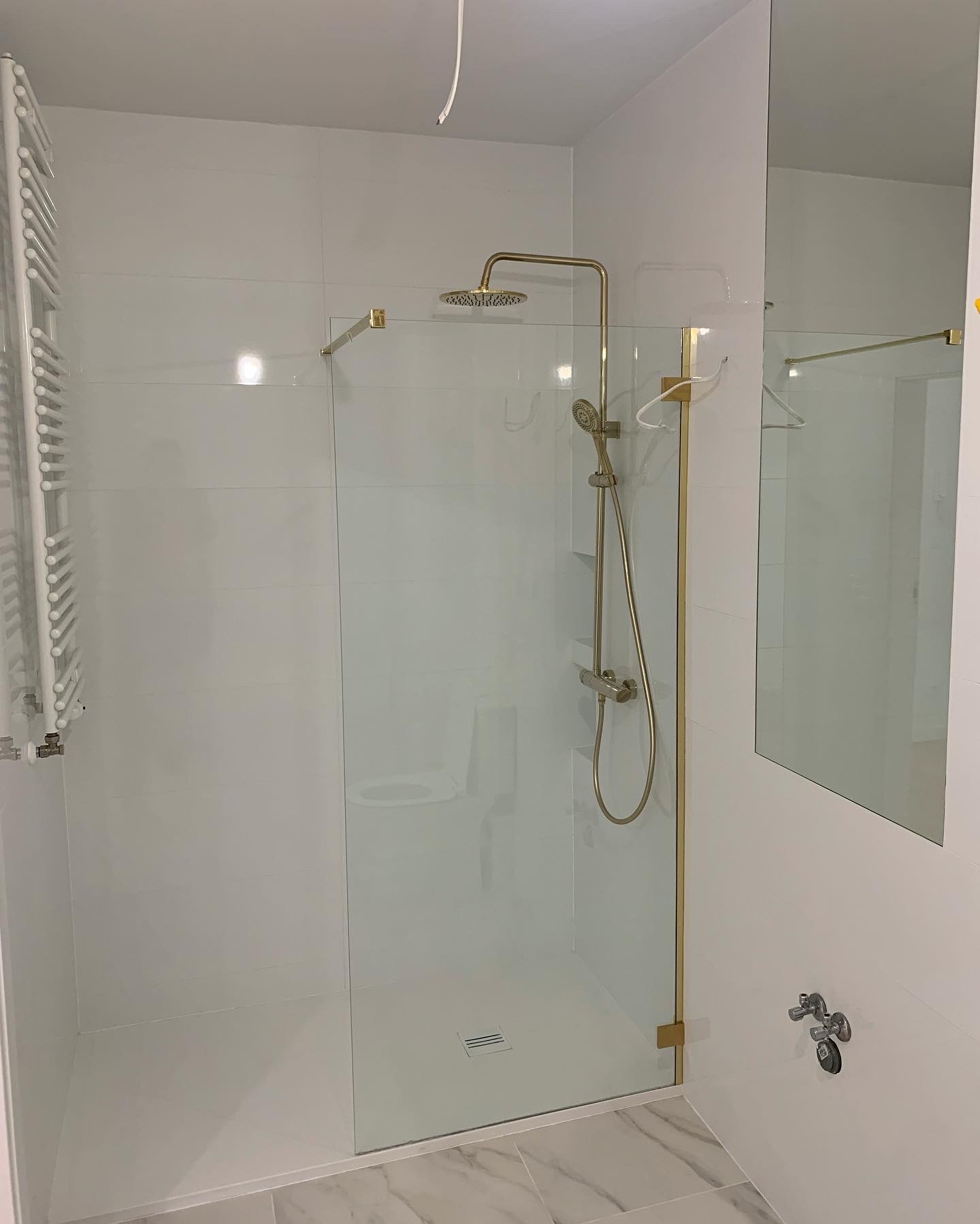 Nowoczesna kabina prysznicowa z elementami w kolorze złotym, białe płytki na ścianach, marmurowa podłoga, widoczny grzejnik drabinkowy i lustro.