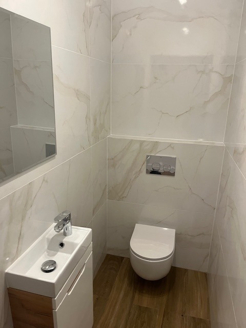 Wykończona łazienka w odcieniach bieli i brązu, z marmurowymi płytkami na ścianach, drewnopodobną podłogą, wiszącą toaletą i małą umywalką z szafką.